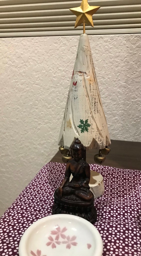A small home altar we have at home, showing a statue of the Buddha sitting beneath a white, miniature Christmas Tree.