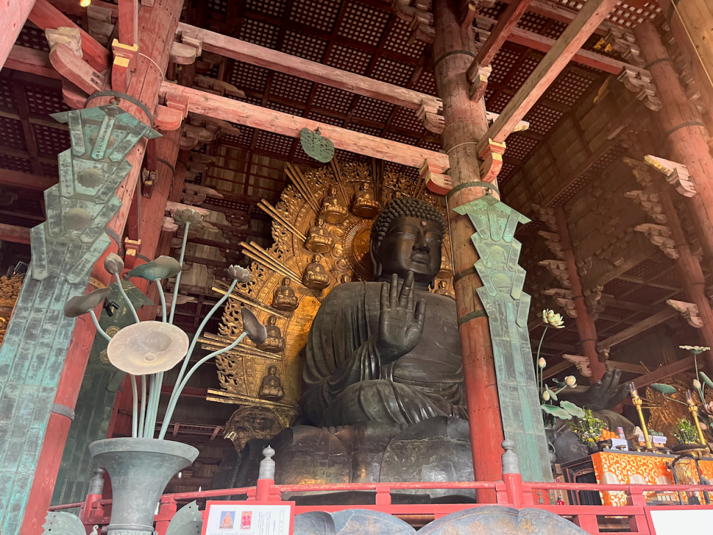 A massive temple interior showing an immense, seated bronze Buddha statue with a similarly massive halo behind it. There are small buddhas in the halo. The buddha holds its palm straight out at the viewer.