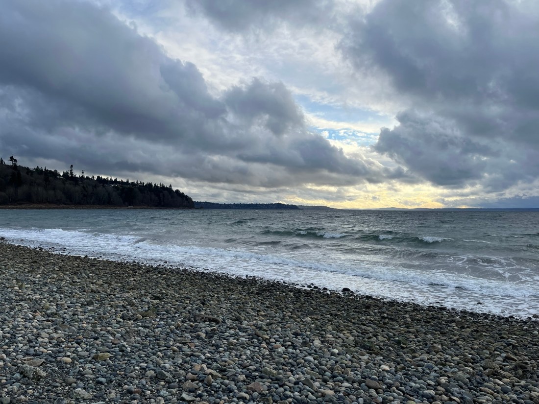 A beach scene with a rocky shore, and waves lapping up to teh shore. In the distance is a cloudy sunset.